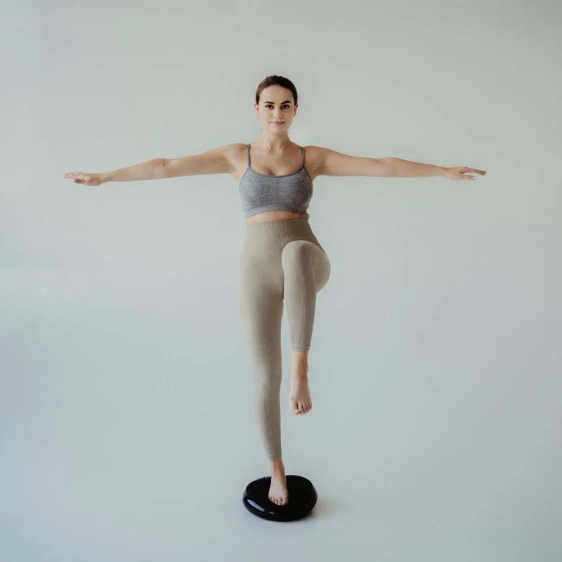 ravnotezni_disk_1 - fizio trgovina woman in a gray sports bra and beige leggings balances on one leg on a black balance disc with arms extended to the sides.