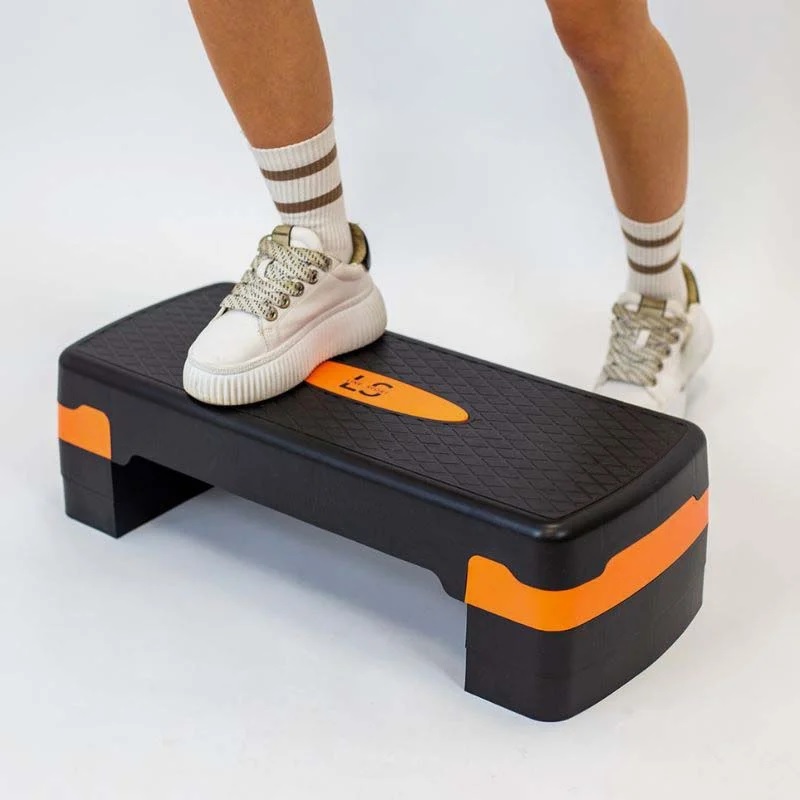 steper_za_aerobiko_11 - fizio trgovina person's foot rests on a black step platform with orange accents during a cardio exercise, wearing white sneakers and striped socks.
