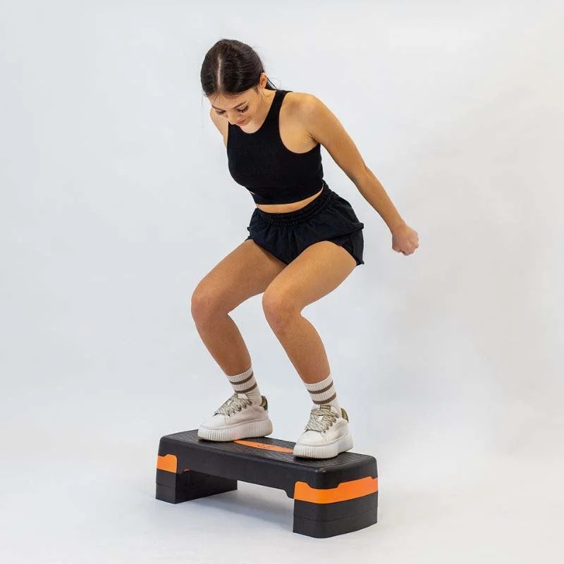steper_za_aerobiko_12 - fizio trgovina woman in a black athletic outfit stepping onto a small black and orange aerobic platform, mid‑exercise.