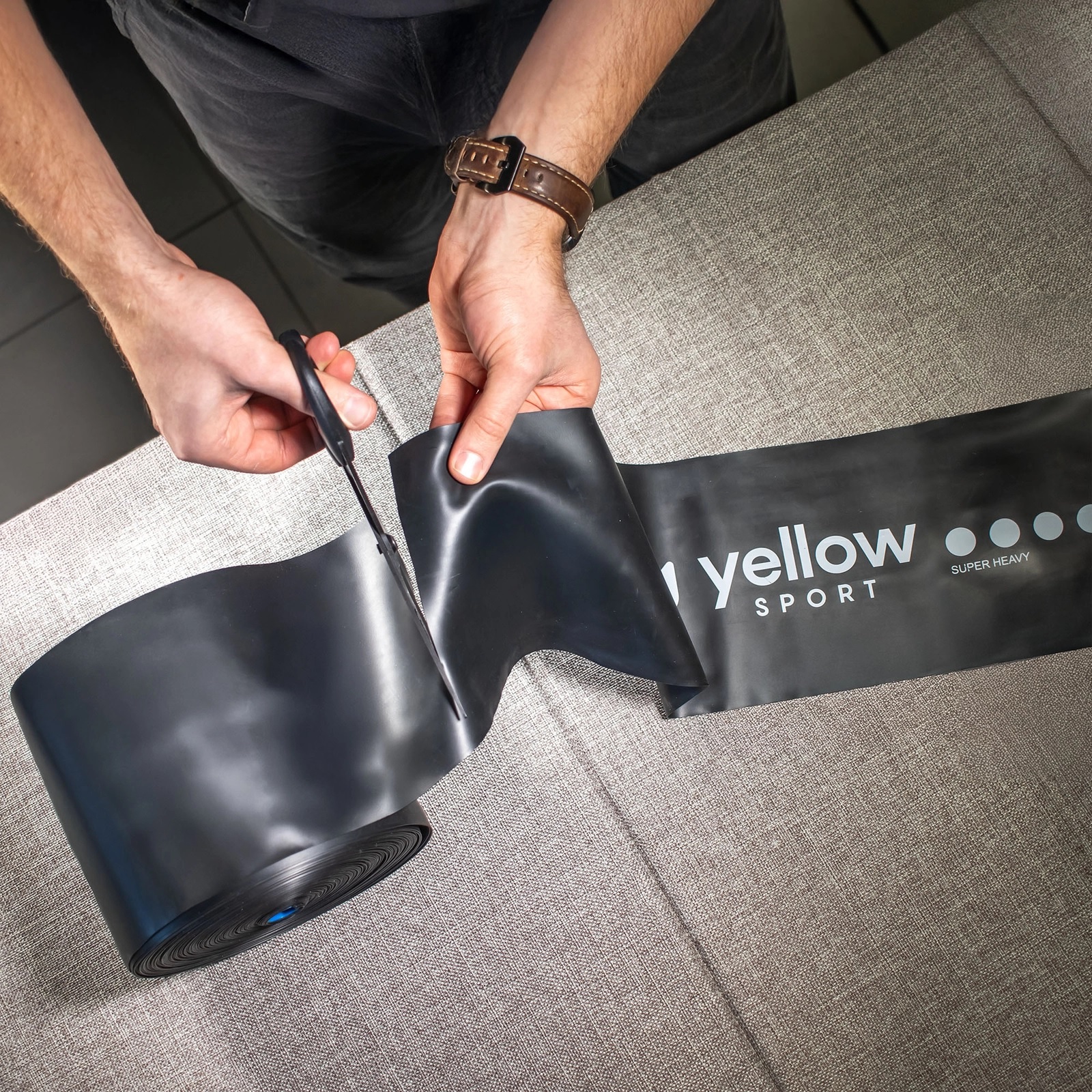 elasticni_trak_30m_28 - fizio trgovina person cuts a rolled black plastic sheet with scissors on a gray fabric surface; brown leather watch visible on wrist, packaging shows 'yellow sport'.