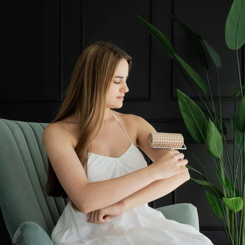 masazni_valjcek_3 - fizio trgovina woman in a white dress sits on a green chair and rollers a skincare dermaroller over her forearm indoors with plants nearby.